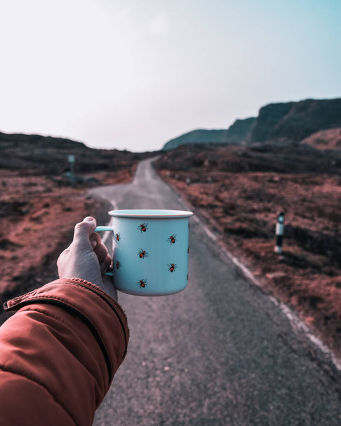 Woman hand holding a coffee mug in the middle of a mountain road - Foto, imagen