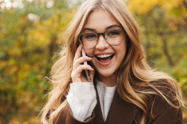 Beautiful young cheerful woman wearing coat walking in the autumn park, talking on mobile phone - Photo, image