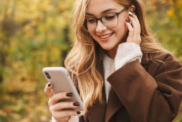 Beautiful young cheerful woman wearing coat walking in the autumn park, wearing wireless earphones, using mobile phone - Foto, immagini