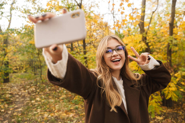 Beautiful young cheerful woman wearing coat walking in the autumn park, taking a selfie - Fotoğraf, Görsel