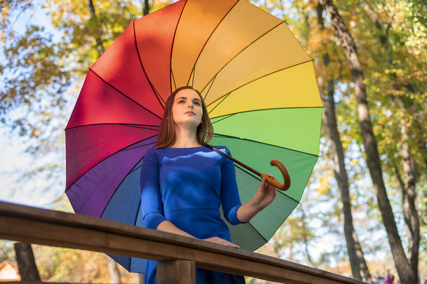 Hermosa joven con el pelo largo en un vestido corto azul con un paraguas de arco iris pasea por el parque de otoño
. - Foto, Imagen