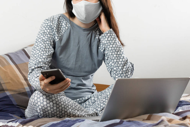 保護マスクの女性はベッドの中で呼び出している。コロナウイルスのパンデミック時の内務省。携帯電話やノートパソコンを使ってオンラインで作業 - 写真・画像