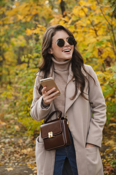 Beautiful young cheerful woman wearing coat walking in the autumn park, using mobile phone - Photo, Image