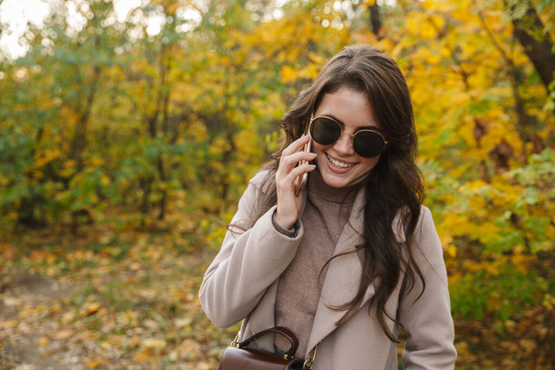 Beautiful young cheerful woman wearing coat walking in the autumn park, talking on mobile phone - Фото, зображення