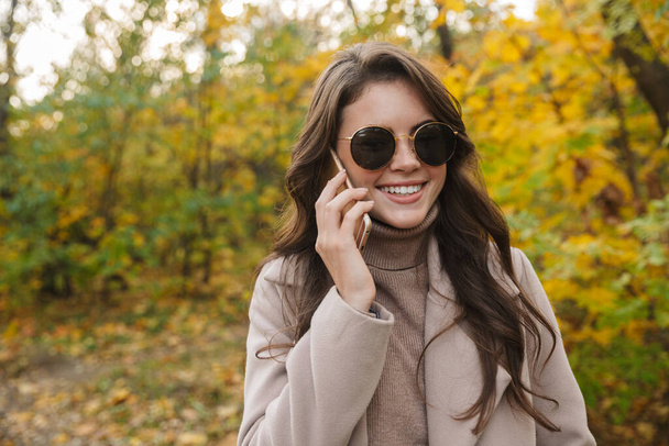 Beautiful young cheerful woman wearing coat walking in the autumn park, talking on mobile phone - Φωτογραφία, εικόνα