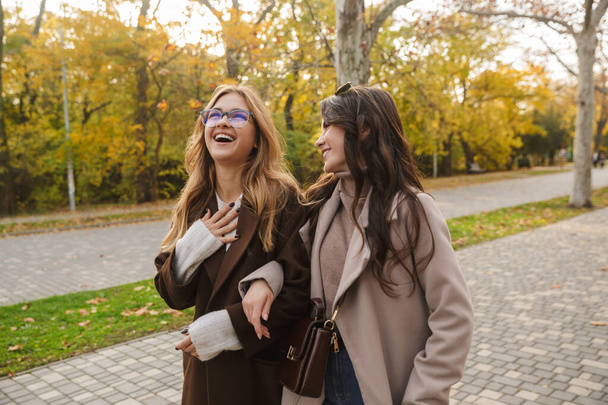 Two cheerful young pretty girls wearing coats walking together at the autumn park - Foto, afbeelding
