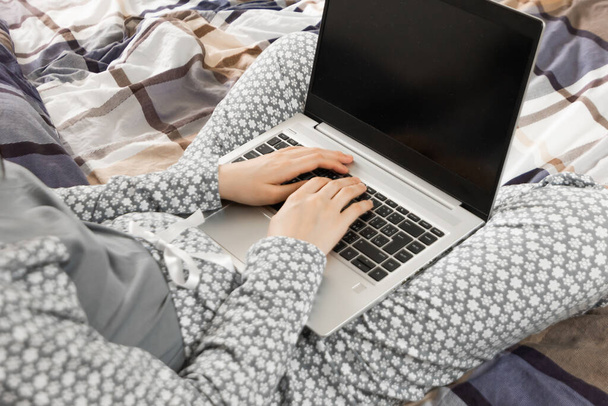 Elölnézet egy laptop képernyőjén egy szürke virágos hálóinges nő térdén. Otthoni iroda koncepció. A Freelancer ágyban dolgozik a coronavirus kitörése alatt. - Fotó, kép