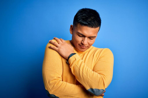 Młody, przystojny Latynos ubrany w żółty, luźny sweter na odizolowanym niebieskim tle, przytulający się radośnie i pozytywnie, z uśmiechem na twarzy. Miłość do siebie i dbanie o siebie - Zdjęcie, obraz