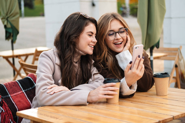Two cheerful attractive women friends sitting at the cafe outdoors, looking at mobile phone while drinking coffee - Fotó, kép