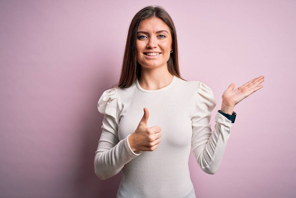 Young beautiful woman with blue eyes wearing casual white t-shirt over pink background Showing palm hand and doing ok gesture with thumbs up, smiling happy and cheerful - Valokuva, kuva