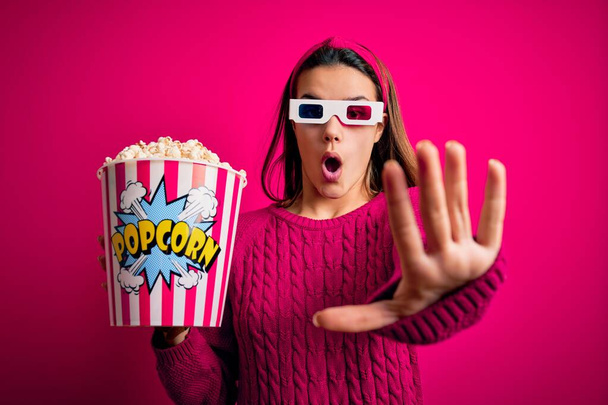Young beautiful girl watching movie using 3d glasses eating box with popcorns doing stop gesture with hands palms, angry and frustration expression - Foto, Imagen