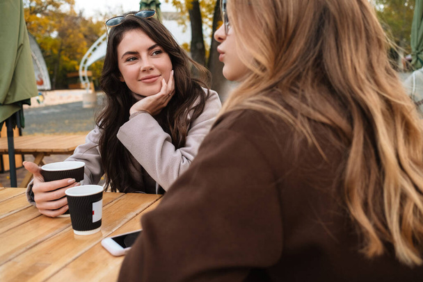 Дві веселі привабливі друзі сидять у кафе на відкритому повітрі, п'ють каву
 - Фото, зображення