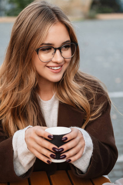 Close up of a smiling young attractive woman wearing autumn coat drinking takeaway coffee outdoors - Φωτογραφία, εικόνα