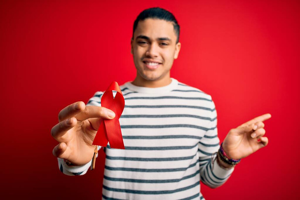 Young brazilian man holding red hiv ribbon standing over isolated background very happy pointing with hand and finger to the side - Φωτογραφία, εικόνα