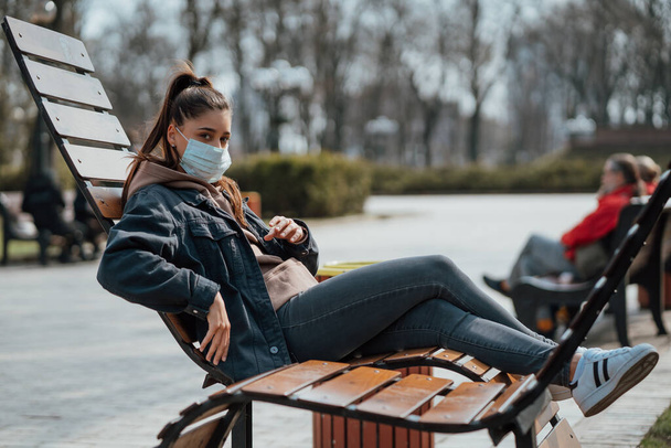Yong femme assise sur le banc dans le parc et portant un masque de protection. Concept de coronavirus
. - Photo, image