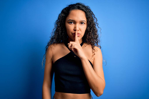 Young beautiful woman with curly hair wearing casual t-shirt standing over blue background asking to be quiet with finger on lips. Silence and secret concept. - Photo, Image