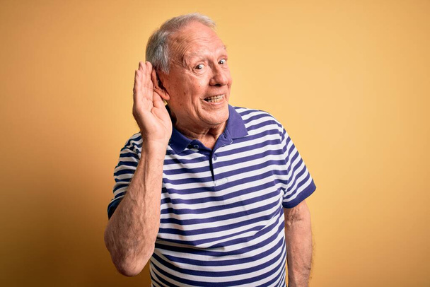 Homem idoso de cabelos grisalhos vestindo camiseta listrada da marinha casual em pé sobre fundo amarelo sorrindo com a mão sobre o ouvido ouvindo uma audiência para boatos ou fofocas. Conceito de surdez
. - Foto, Imagem
