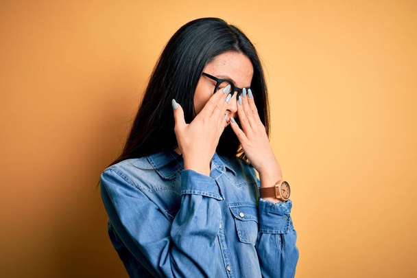 Jovem mulher chinesa bonita vestindo camisa jeans casual sobre fundo amarelo isolado esfregando os olhos para fadiga e dor de cabeça, sonolento e expressão cansada. Problema de visão
 - Foto, Imagem