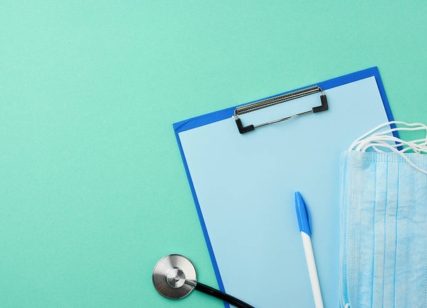 металлический стетоскоп, держатель бумаги с чистым голубым листом, одноразовая синяя медицинская маска на зеленом фоне, вид сверху, плоский
 - Фото, изображение