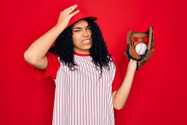 Fiatal afro-amerikai göndör sportos nő sapkában baseballozik labdával és kesztyűvel a fején, szégyenkezve és meglepett arccal, dühös és frusztrált. Félelem és tévedés. - Fotó, kép