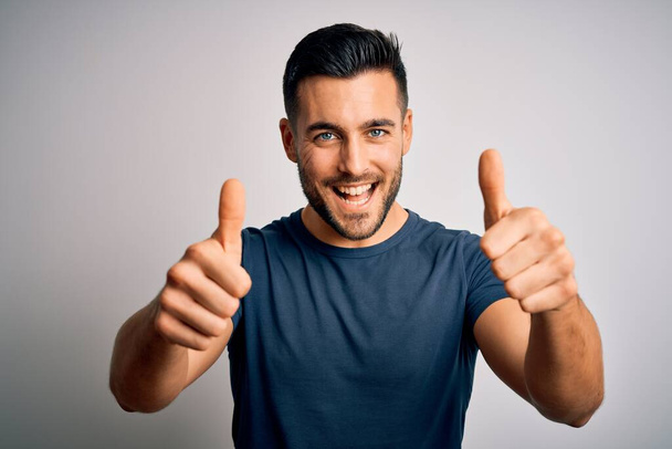 Young handsome man wearing casual t-shirt standing over isolated white background approving doing positive gesture with hand, thumbs up smiling and happy for success. Winner gesture. - Photo, Image