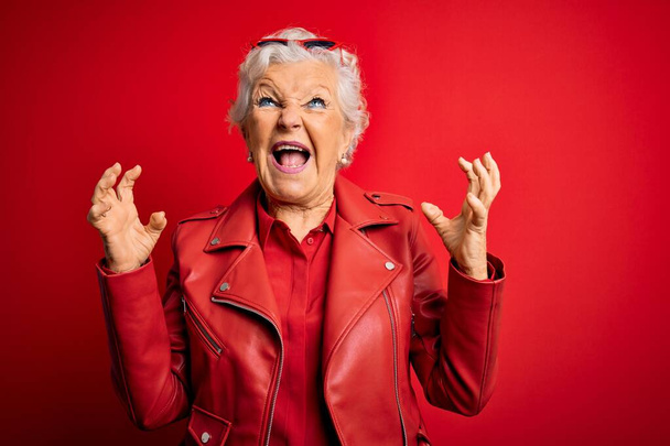 Senior beautiful grey-haired woman wearing casual red jacket and sunglasses crazy and mad shouting and yelling with aggressive expression and arms raised. Frustration concept. - Zdjęcie, obraz