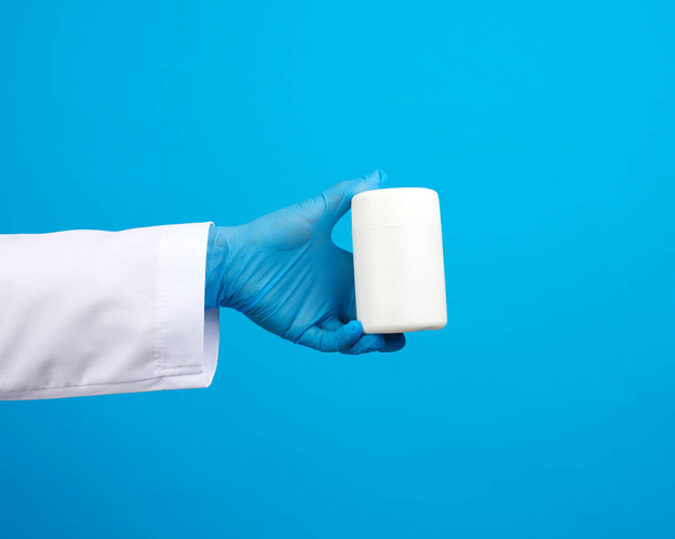 kéz a kék steril kesztyű tart egy fehér műanyag edény tabletták, orvosi kezelés koncepció betegségek, kék háttér - Fotó, kép
