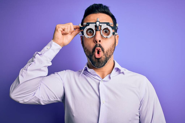 Jonge knappe optische man met baard dragen optometrie bril over paarse achtergrond bang in shock met een verrassing gezicht, bang en opgewonden met angst uitdrukking - Foto, afbeelding