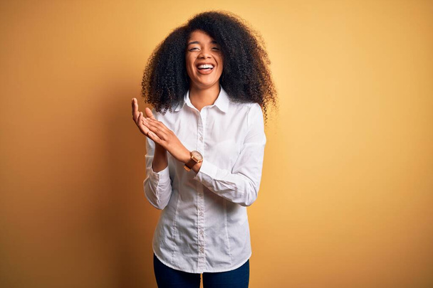 Fiatal gyönyörű afro-amerikai elegáns nő afro haj áll sárga háttér tapsol és tapsol boldog és örömteli, mosolygós büszke kezek együtt - Fotó, kép