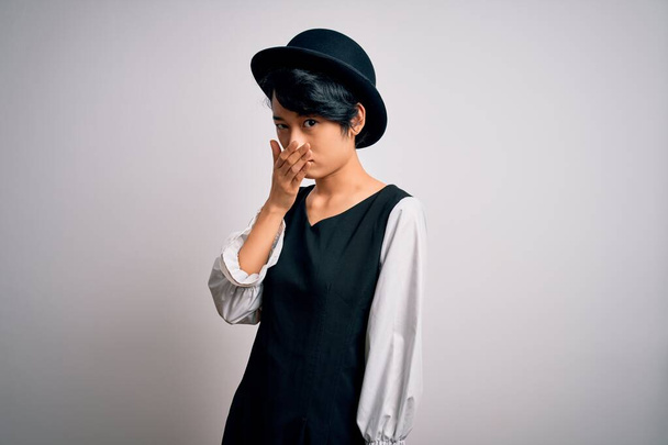Young beautiful asian girl wearing casual dress and hat standing over isolated white background smelling something stinky and disgusting, intolerable smell, holding breath with fingers on nose. Bad smell - Photo, Image
