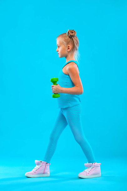 Παιδί κορίτσι κάνει ασκήσεις φυσικής κατάστασης ή γιόγκα με αλτήρες απομονώνονται σε μπλε φόντο - Φωτογραφία, εικόνα