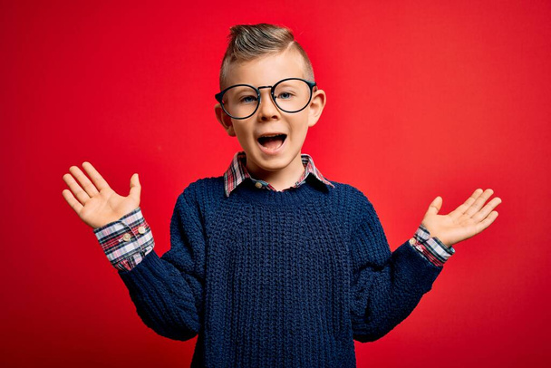 Junge kleine kaukasische Kinder mit blauen Augen stehen mit smarter Brille vor rotem Hintergrund und feiern verrückt und staunen über den Erfolg mit erhobenen Armen und offenen Augen, die aufgeregt schreien. Siegerkonzept - Foto, Bild