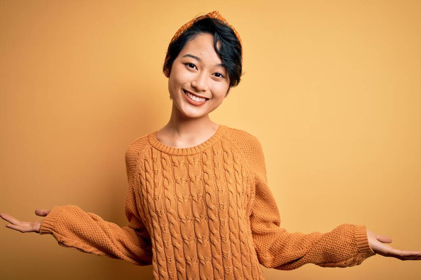 Fiatal gyönyörű ázsiai lány visel alkalmi pulóver és diadem áll sárga háttér mosoly mutatja mindkét kezét nyitott tenyér, bemutató és reklám összehasonlítás és az egyensúly - Fotó, kép