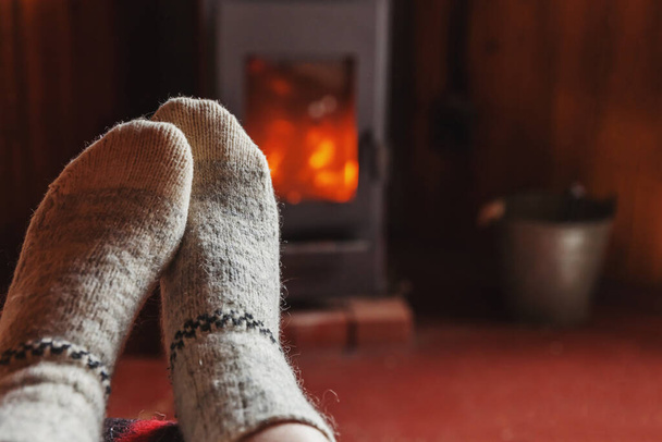 Pies piernas en ropa de invierno calcetines de lana en el fondo de la chimenea. Mujer sentada en casa en invierno u otoño por la noche relajándose y calentándose. Concepto de invierno y clima frío. Hygge Nochebuena - Foto, Imagen
