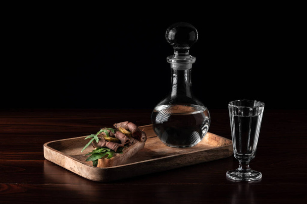 Crystal decanter or carafe with a glass full of vodka and tasty appetizer bruschetta with bacon and pickles on wooden board, alcohol drink concept with copy space - Zdjęcie, obraz
