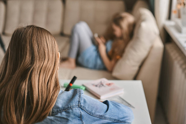 Uma adolescente faz seu dever de casa enquanto sua irmã joga em seu telefone. Comunicação, distância social durante a quarentena. Ensino à distância, educação online
 - Foto, Imagem