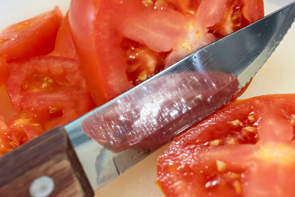 Vista de perto de uma faca de bife com uma pega de madeira cortando através de um tomate de bife vermelho em uma placa de corte
. - Foto, Imagem