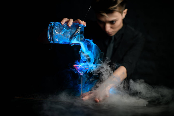 female barman professionally pours illuminated smoky cocktail from mixing bowl into martini glass. - Foto, imagen