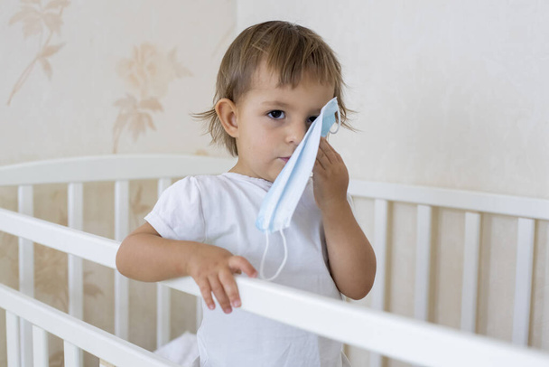 概念コロナウイルスは隔離を冷却します。かわいい幼児は家のベビーベッドに立って医療マスクを抱いています - 写真・画像
