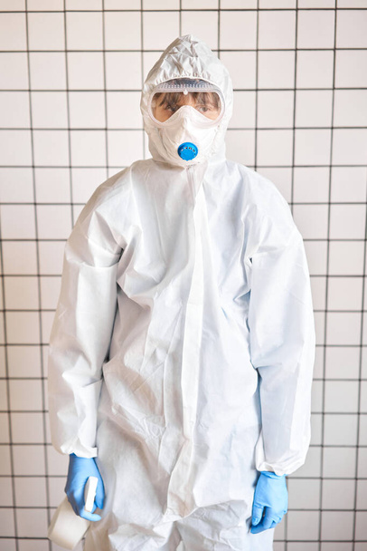 Beyaz sprey şişesi koruyucu bir takım elbise ve mavi eldivenli kadınların ellerinde. Dezenfeksiyon, temizlik ve yıkama. COVID-19. Koronavirüs enfeksiyonunun önlenmesi. - Fotoğraf, Görsel