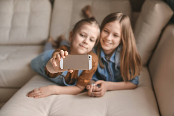 Beautiful girls take selfies on their phone while lying on the sofa. Sisters take a break from homework and take photos during quarantine - Photo, Image