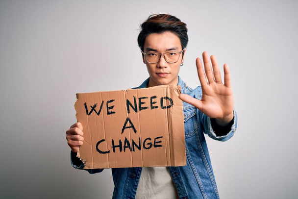 Fiatal jóképű kínai aktivista tiltakozó kéri változás tartó poszter nyitott kézzel csinál stop jel komoly és magabiztos kifejezés, védelmi gesztus - Fotó, kép