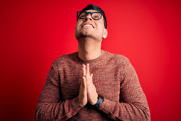 Homem hispânico bonito jovem vestindo óculos nerd sobre fundo vermelho implorando e orando com as mãos, juntamente com a expressão de esperança no rosto muito emocional e preocupado. Implorando
. - Foto, Imagem