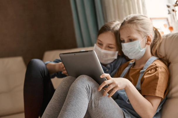 Két csinos lány orvosi maszkban ül a kanapén egy tablettával. Az iskoláslányok megcsinálják a házi feladatukat. Távolságtanulás a COVID-19 karantén alatt. - Fotó, kép