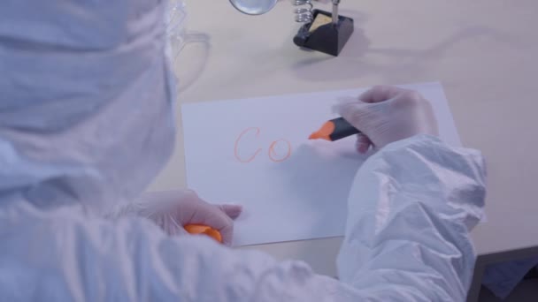 Health care worker in protective antiviral suit writing Covid-19 on a sheet of paper. Global pandemic problem, coronavirus infectious disease, lockdown, quarantine. Shooting over shoulder. - Felvétel, videó