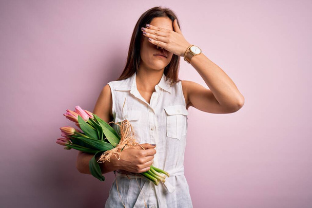 Jeune belle femme brune tenant bouquet de fleurs de tulipes sur fond rose couvrant les yeux avec la main, l'air grave et triste. Concept d'appréhension, de dissimulation et de rejet
 - Photo, image