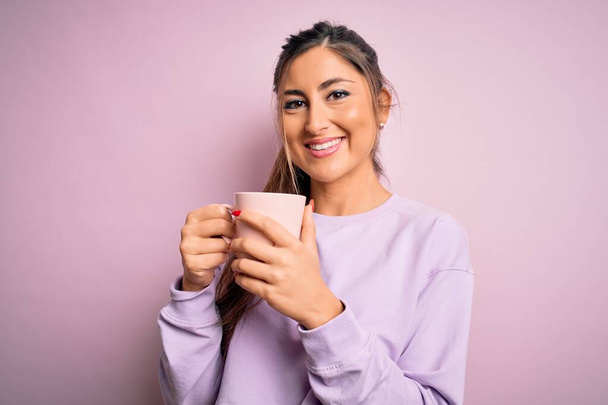 Νεαρή όμορφη μελαχρινή γυναίκα πίνοντας κούπα καφέ πάνω από απομονωμένο ροζ φόντο με ένα χαρούμενο πρόσωπο στέκεται και χαμογελά με αυτοπεποίθηση χαμόγελο που δείχνει τα δόντια - Φωτογραφία, εικόνα