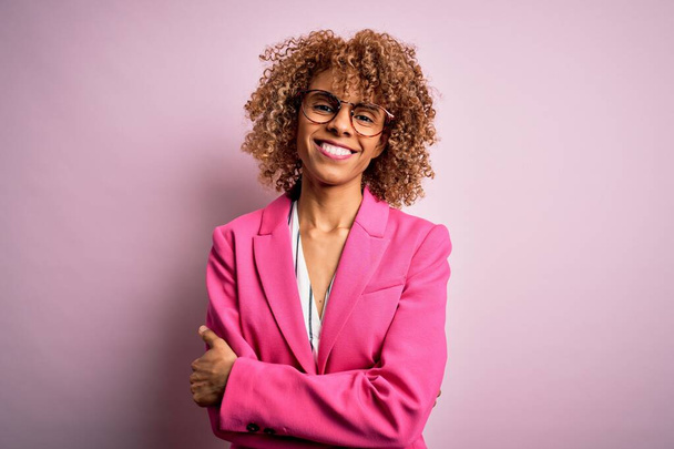 Młoda afrykańska bizneswoman w okularach stojąca nad różowym tłem szczęśliwa twarz uśmiechnięta skrzyżowanymi ramionami patrząca w kamerę. Osoba pozytywna. - Zdjęcie, obraz