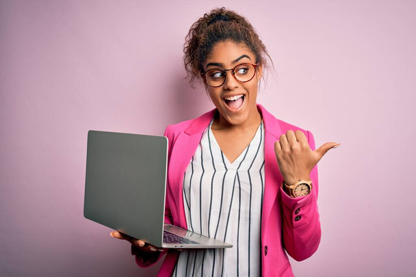 Afroamerykanka biznes kobieta noszenie okularów pracy przy użyciu laptopa na różowym tle wskazując i pokazując z kciukiem do góry z uśmiechem na twarzy szczęśliwy - Zdjęcie, obraz