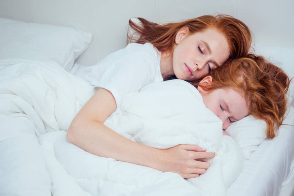 hermosa pelirroja pelirroja mujer abrazando dormido su hija bajo manta blanca en acogedora pandemia de infección home.corona mañana
 - Foto, Imagen